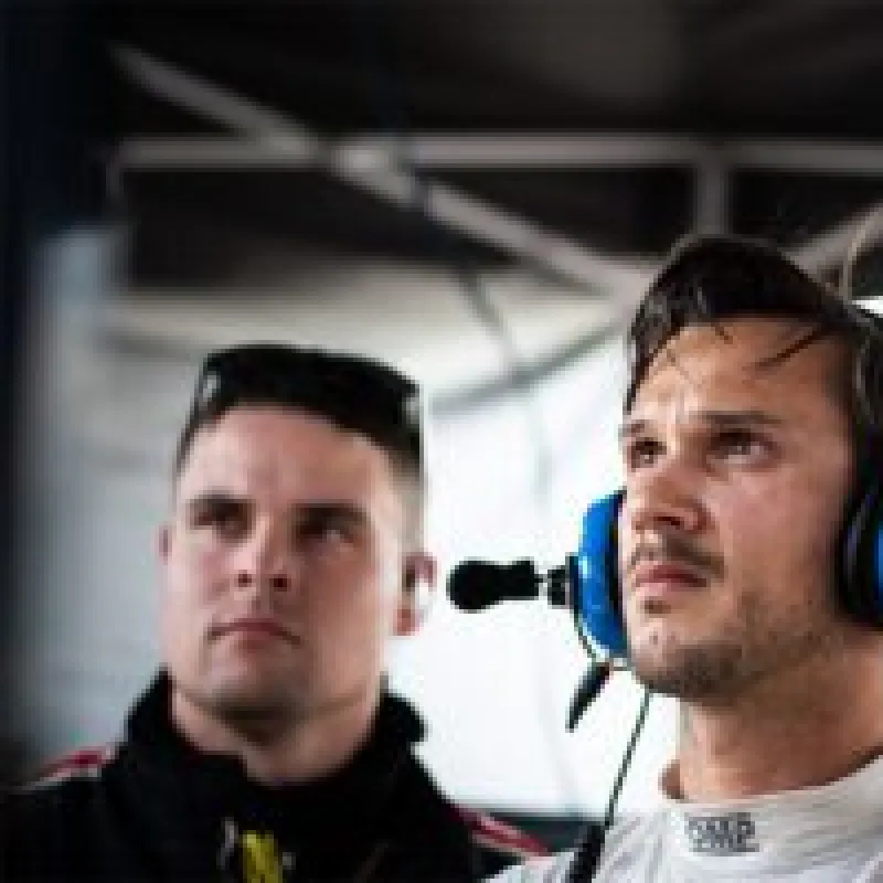 Two men focused at a racing event, one wearing a headset with microphone and racing gear in a pit area.