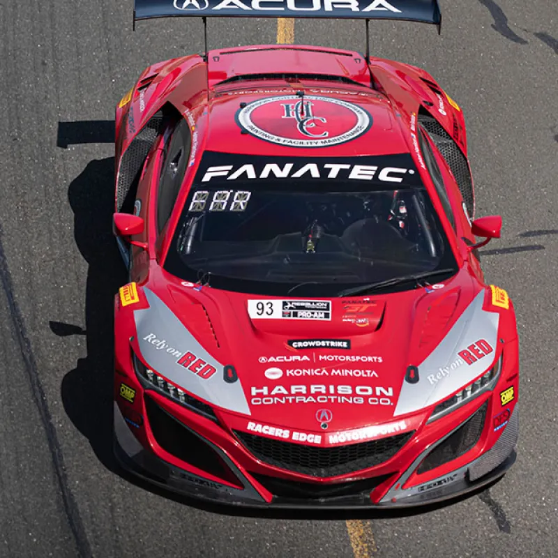 Red Acura race car with Fanatec and Harrison Contracting logos on racetrack with asphalt and yellow line.