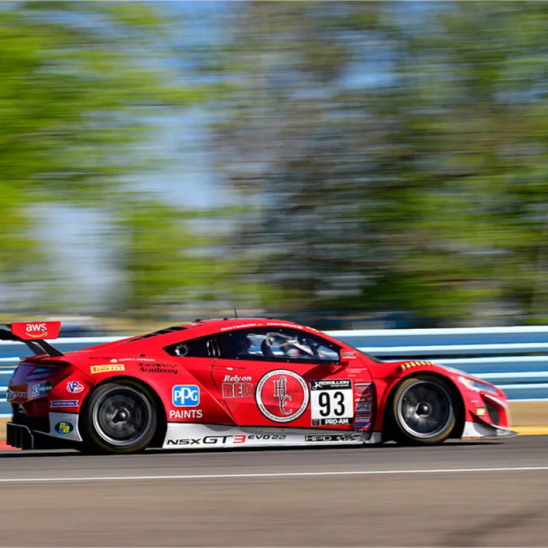 Red NSX GT3 Evo race car number 93 speeding on race track with blurred green background and blue barriers.