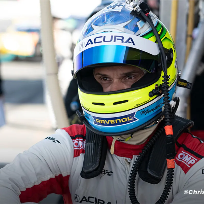 Race car driver in Acura helmet and suit prepares in the pit area during motorsport event.