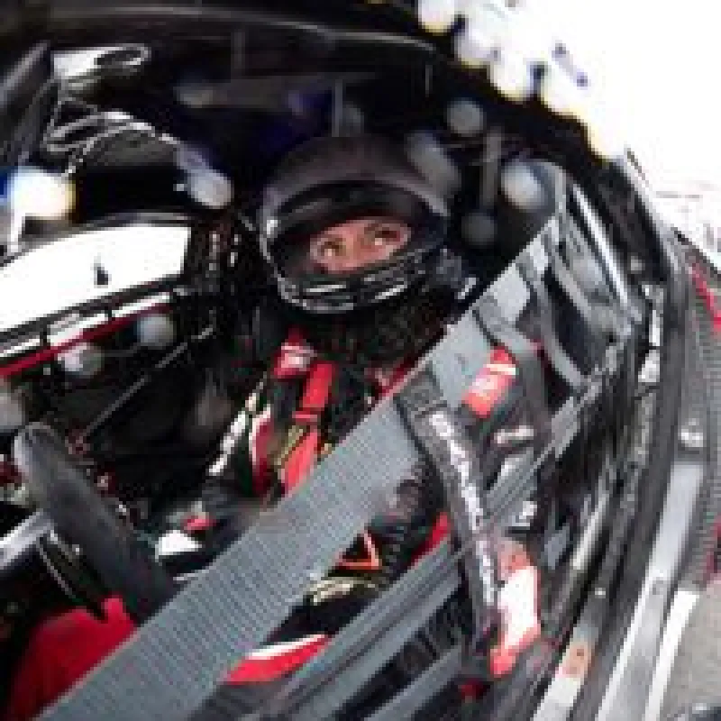 Race car driver wearing a helmet and safety harness inside a racing car cockpit.
