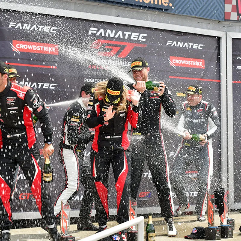 Race car drivers celebrating on podium spraying champagne after GT World Challenge race victory at Watkins Glen.