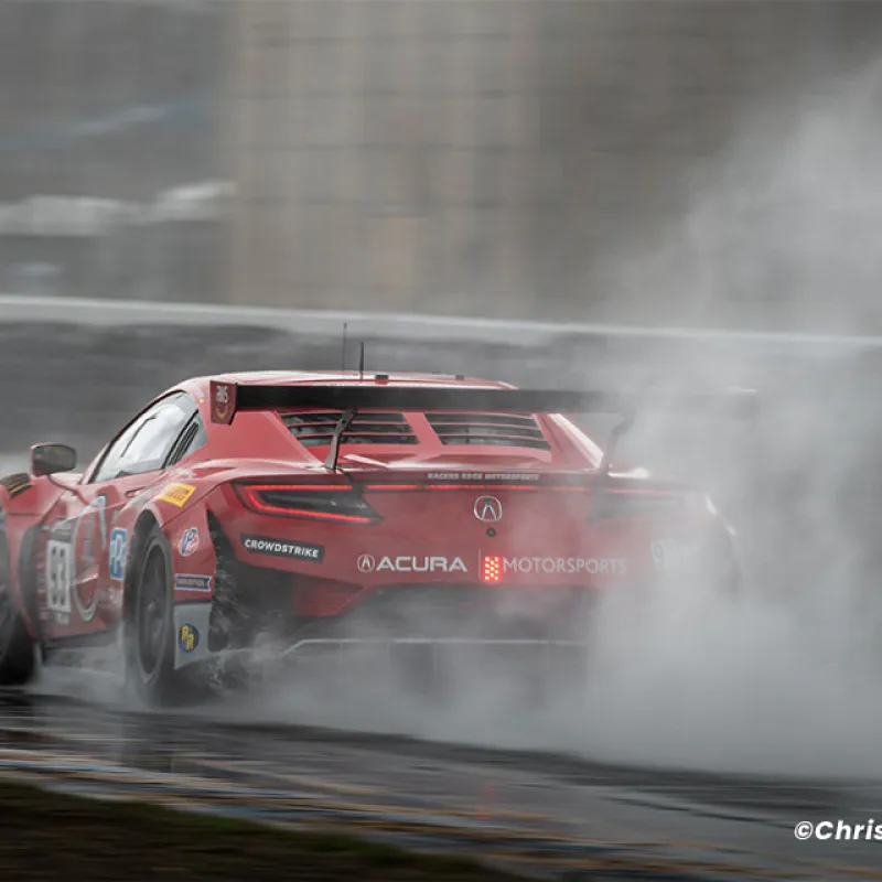 Red Acura race car speeding on wet track, kicking up large spray of water during motorsport event.