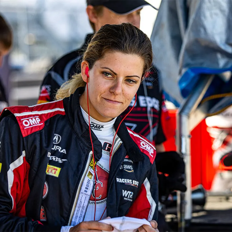 Female race car driver in branded racing suit with earphones, preparing in the pit area before a race.