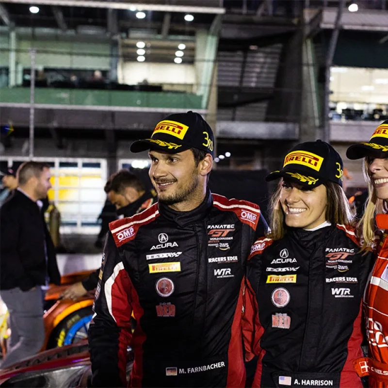 Three smiling race car drivers in racing suits and Pirelli 3rd place hats posing at a nighttime event