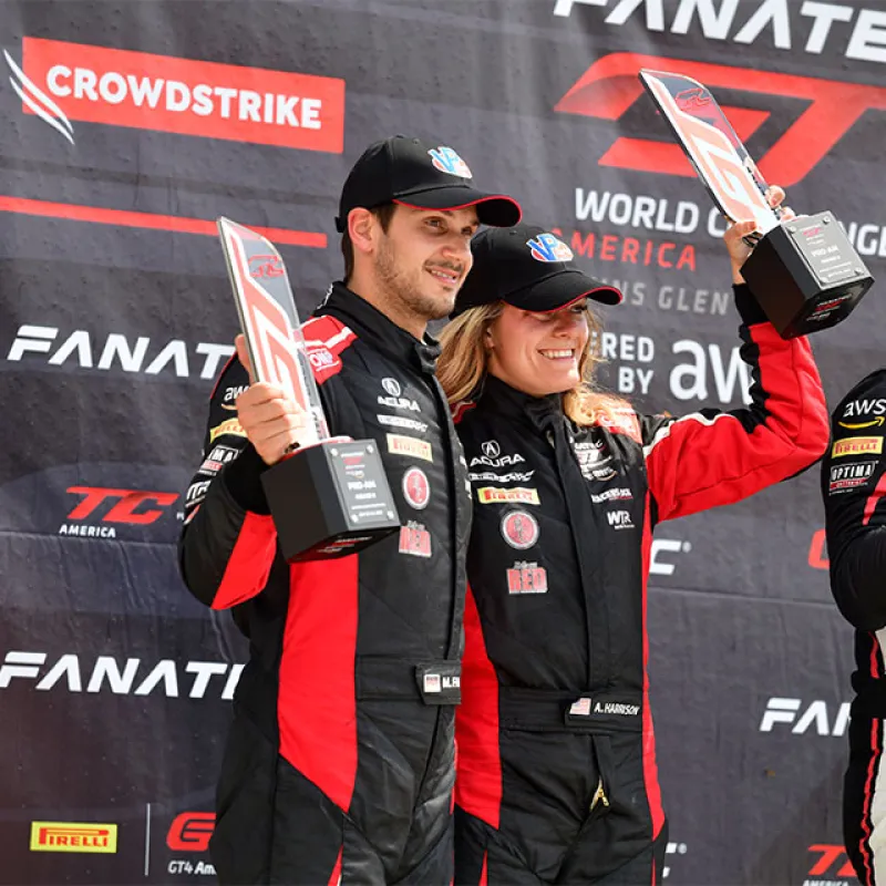Race car drivers in black and red suits hold trophies on podium celebrating their win at GT World Challenge America.