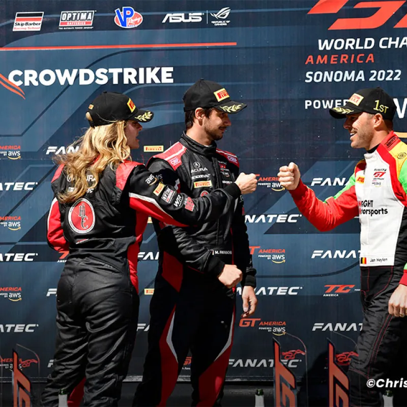 Three race drivers in racing suits and caps fist bump on the GT World Challenge America Sonoma 2022 podium.