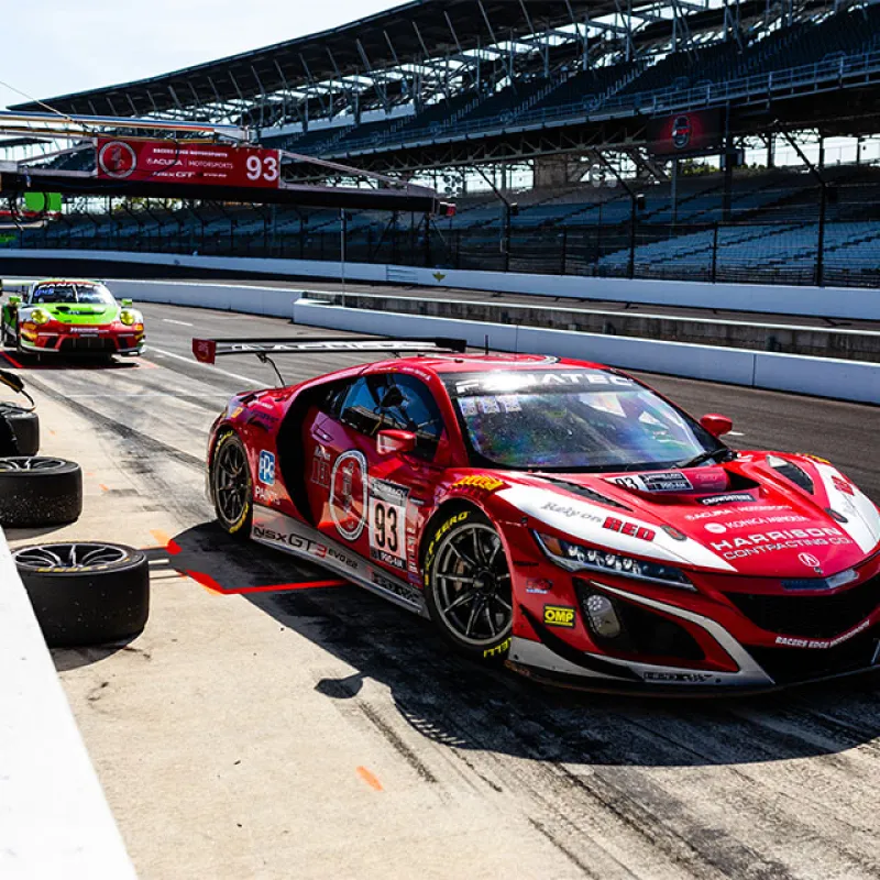Red and green racing cars in a pit stop area with crew members at a professional race track.