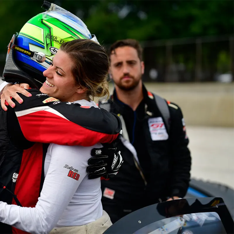 Race car driver in helmet hugging a smiling woman in racing suit near a blue race car with a bearded man in background