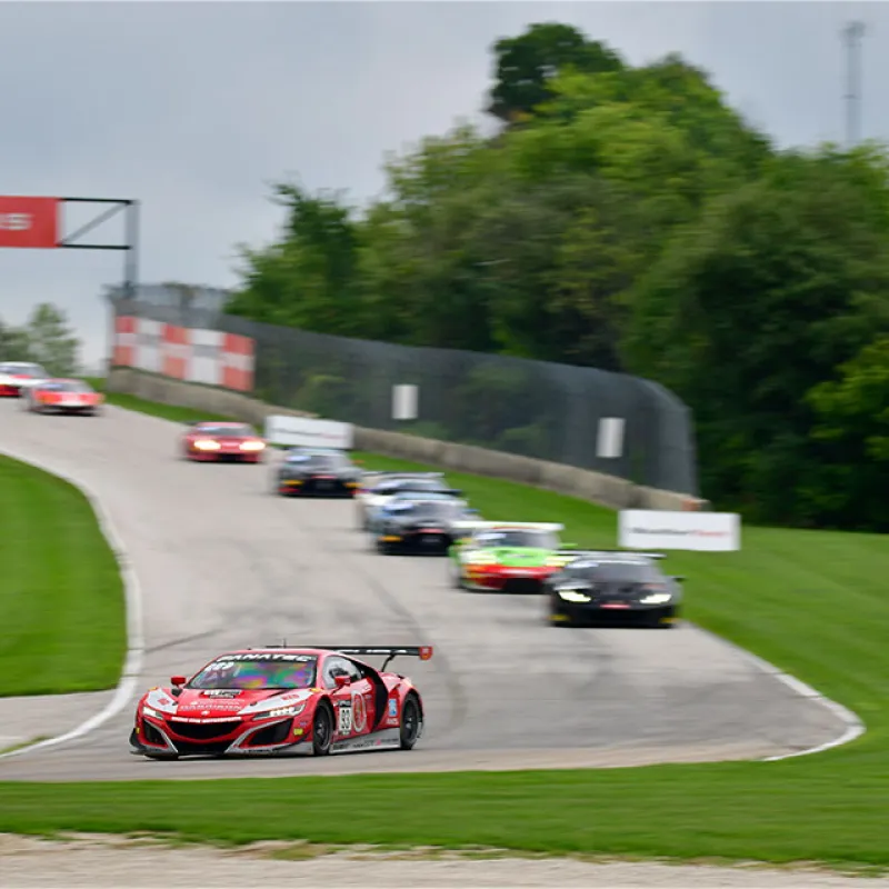 Red racing car leads a pack of sports cars on a curving race track surrounded by green grass and trees.