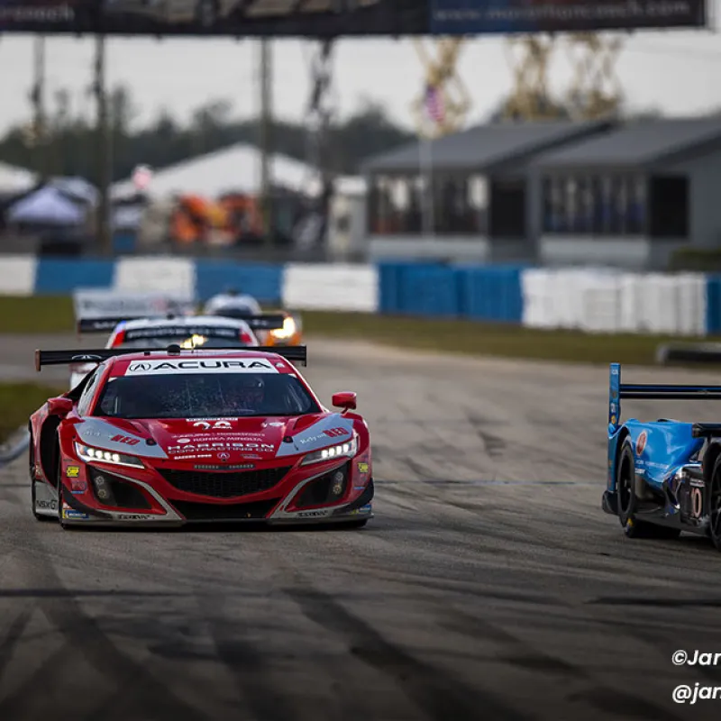 Red and blue Acura race cars competing on a racetrack with blurred background and tire marks on the road.
