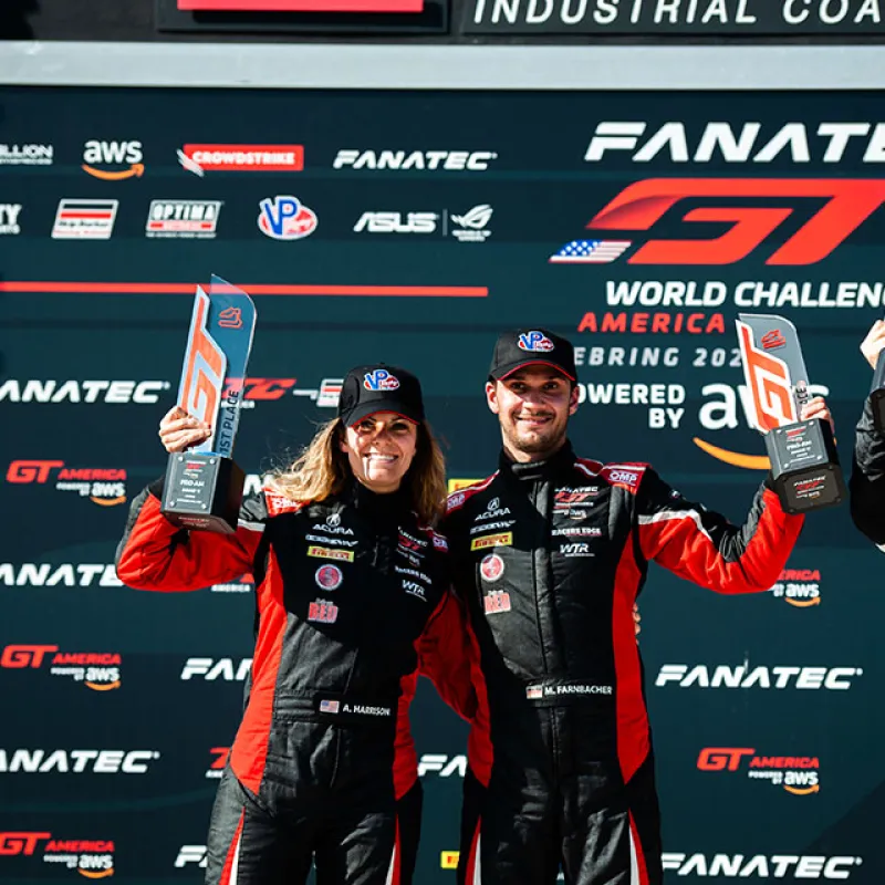Two race car drivers in black and red suits holding trophies on GT America motorsports podium with sponsor logos.