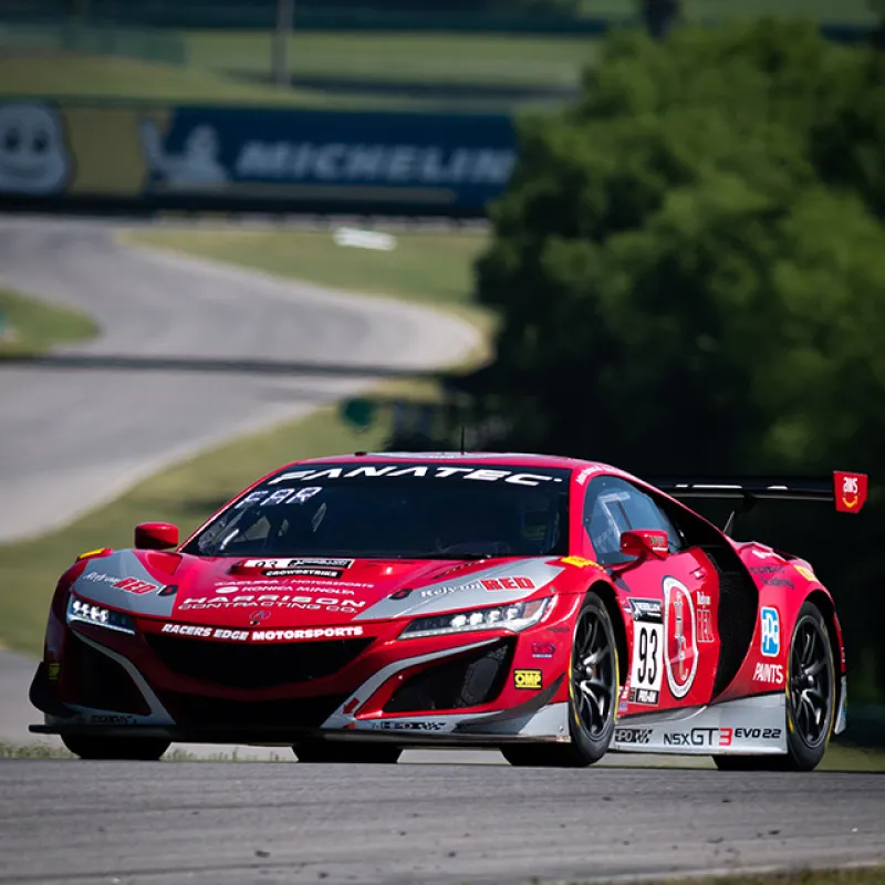 Red race car number 93 speeding on racetrack with Michelin banner and green trees in background.