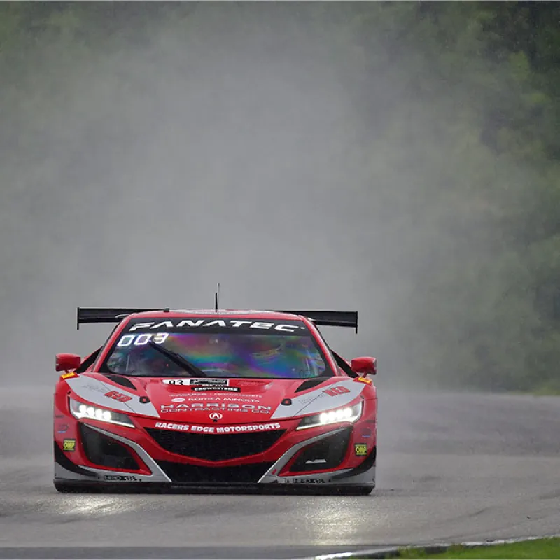 Red Acura NSX race car speeding on wet track with headlights on and mist trailing behind.