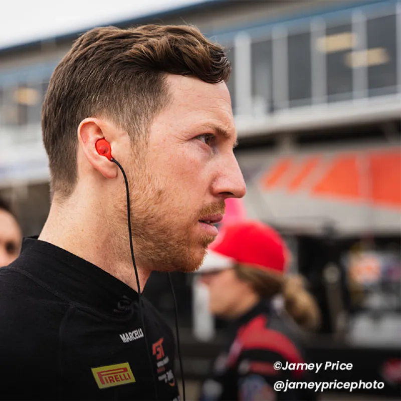Side profile of a focused male race driver wearing earphones in a racing pit area with team members blurred background.