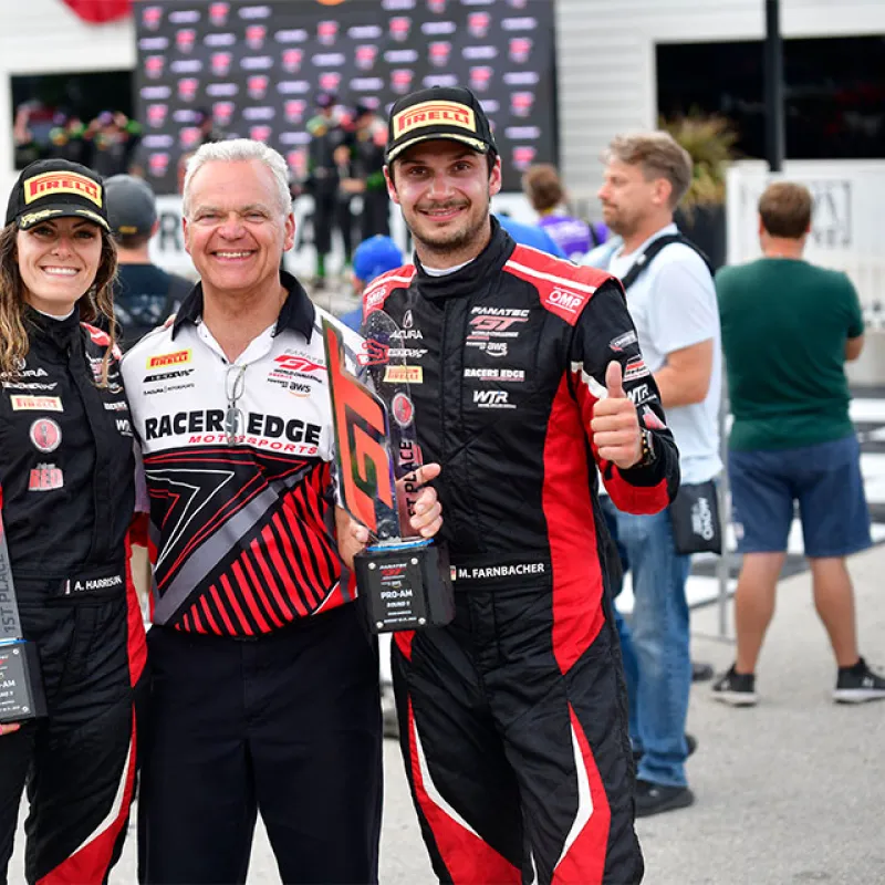 Two racecar drivers holding trophies with a smiling man in the center, all in racing gear at a motorsport event.