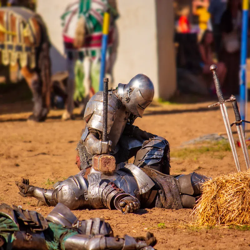 Two knights in armor reenacting a medieval battle scene with swords and hammer on sandy ground outdoors.