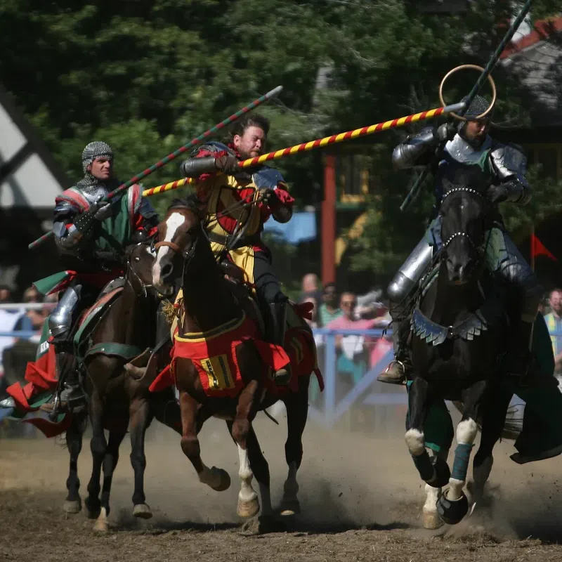 Three armored knights jousting on horses at a medieval festival with crowd and banners in the background.