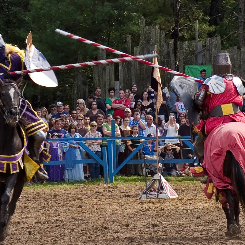 Two armored knights jousting on horseback in front of a lively crowd at a medieval reenactment event.