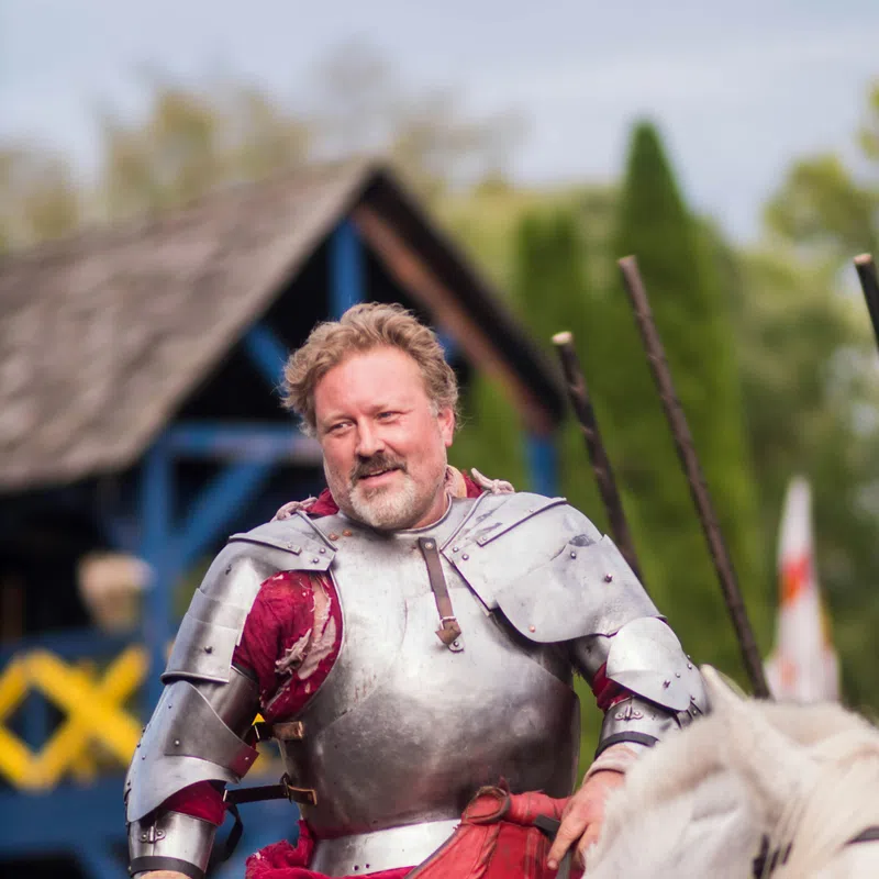 Man in medieval armor riding a white horse at a historical reenactment event with a wooden structure in the background