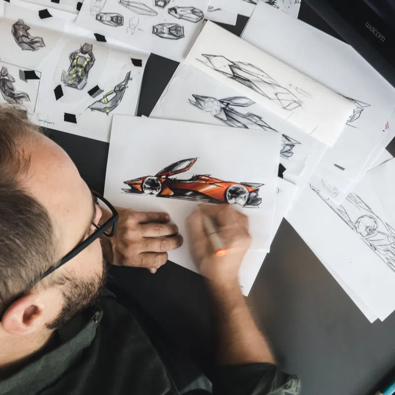 Designer sketching futuristic orange sports car concepts amid multiple design drawings on desk.