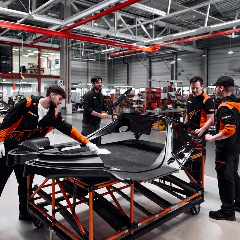 Technicians assemble a black sports car body in a modern factory with industrial equipment and bright lighting.