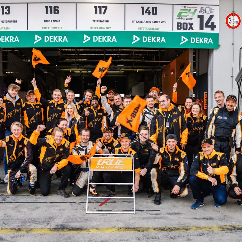 True Racing team posing in front of their garage with orange flags and racing suits at Nürburgring pit lane.