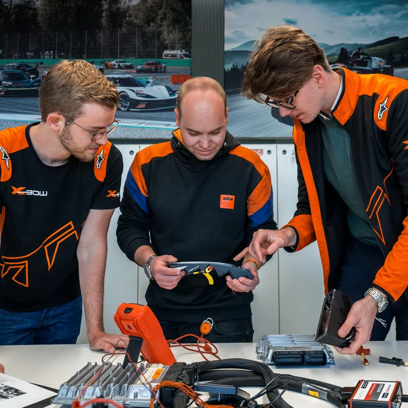 Three engineers in orange and black team gear examining electronic components on a table in a tech workspace.
