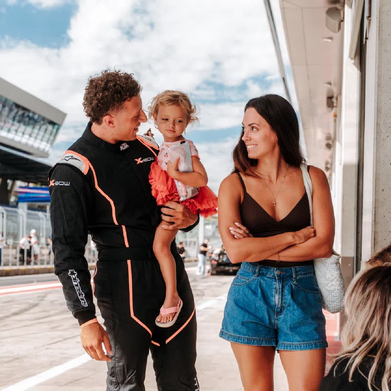 Race car driver in black suit holding a toddler, smiling woman in shorts nearby, and a seated person holding water bottle.