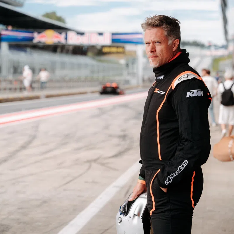 Race car driver in black suit with orange stripes holding helmet, standing on pit lane near Red Bull track signage.
