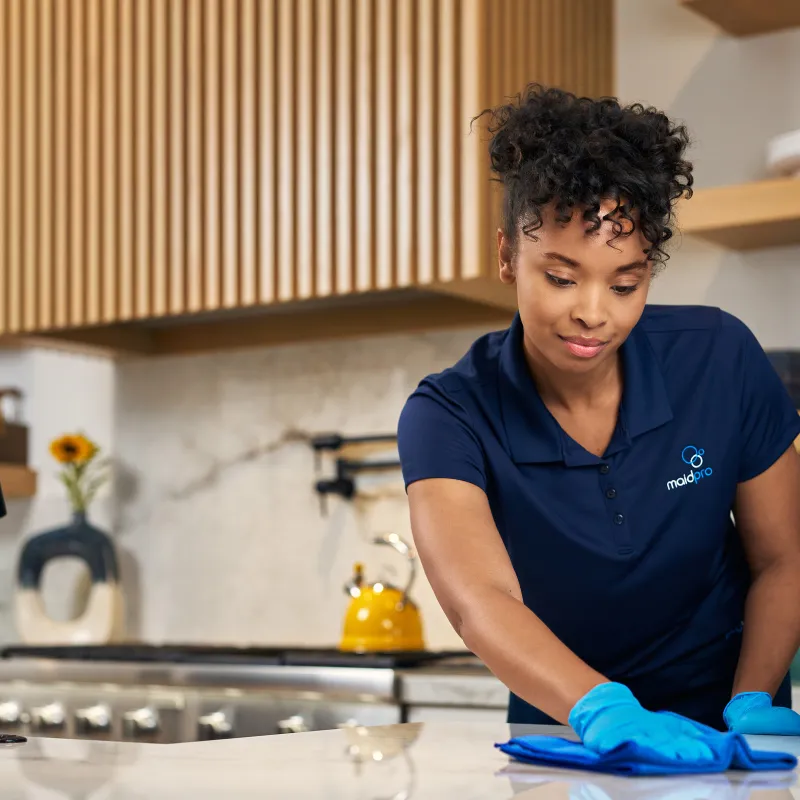 Professional cleaner in blue gloves wiping kitchen countertop with cloth, spray bottle nearby in modern kitchen.