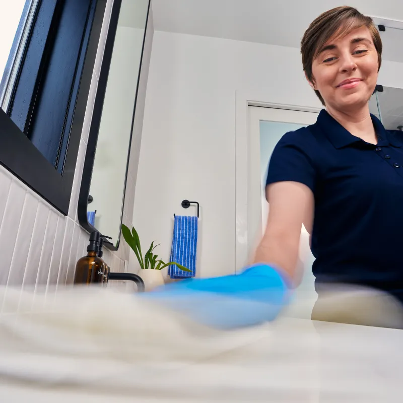 Smiling woman wearing blue gloves cleans a white bathroom sink with a spray bottle and cloth.