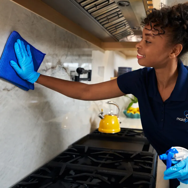 Professional cleaner wearing blue gloves wiping kitchen backsplash with a blue cloth near stove and yellow kettle