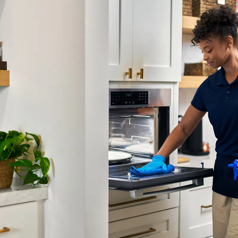 Woman wearing gloves cleaning an open oven door with spray bottle in a modern kitchen.