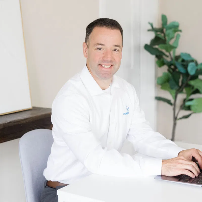 Smiling man in white shirt typing on laptop at white desk in bright modern office with plant and artwork.
