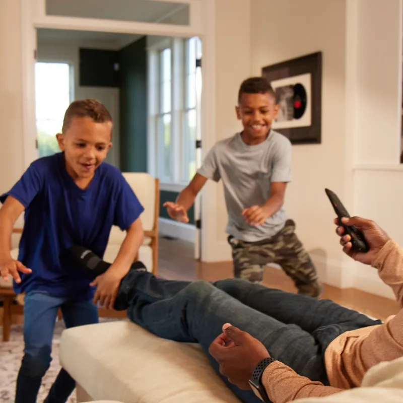 Father relaxing on couch with remote as two young boys playfully run around in living room.