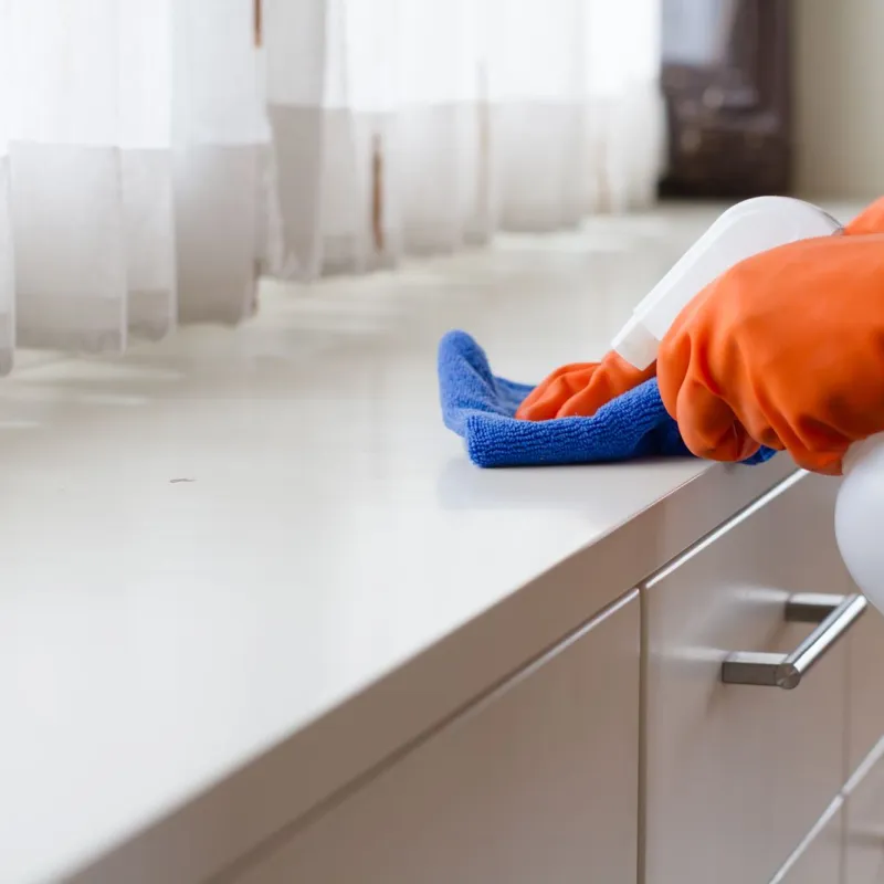 Person wearing orange gloves cleaning a white countertop with a blue cloth and spray bottle near sheer curtains.