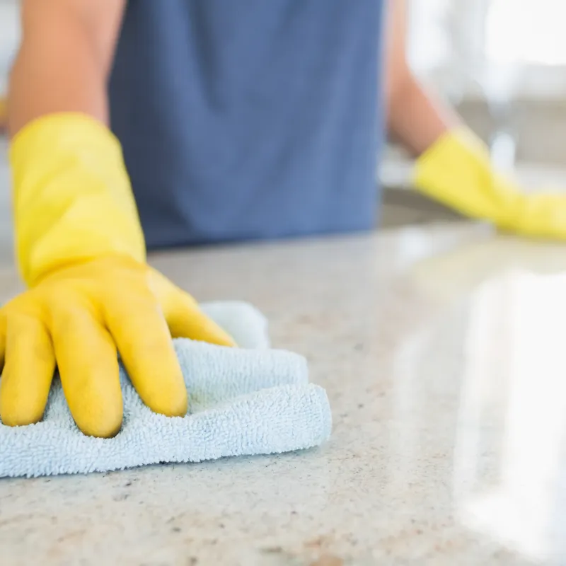 Person wearing yellow gloves cleaning a granite countertop with a light blue microfiber cloth in a bright kitchen.