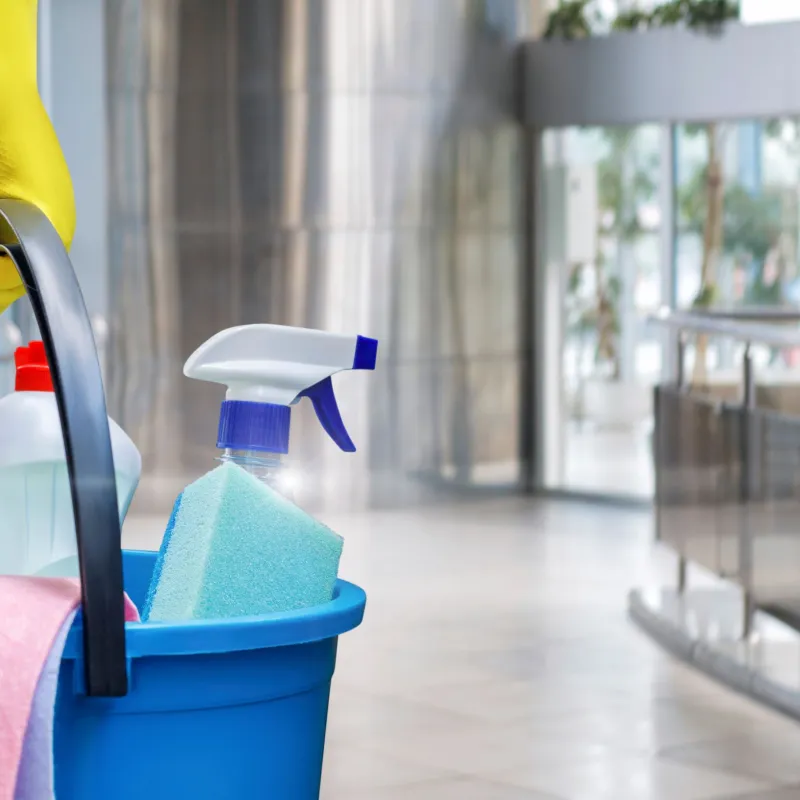 Hand with yellow glove holding blue bucket with cleaning spray, sponge, and supplies in bright modern lobby.