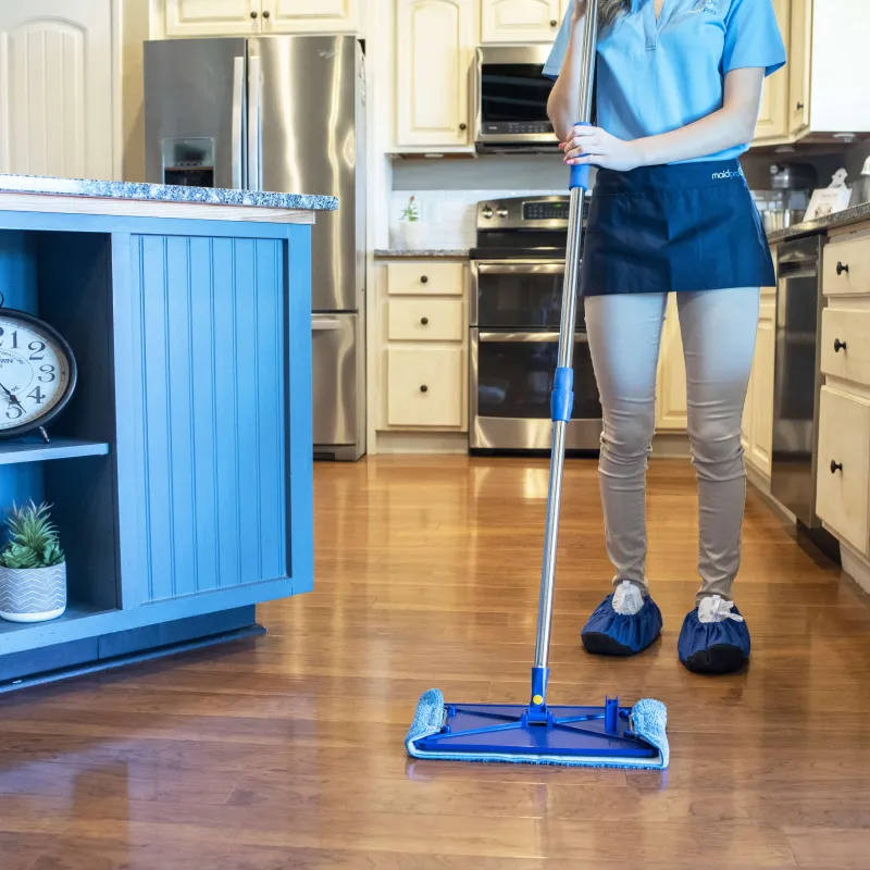 Person mopping hardwood kitchen floor wearing cleaning gear with blue mop and shoe covers