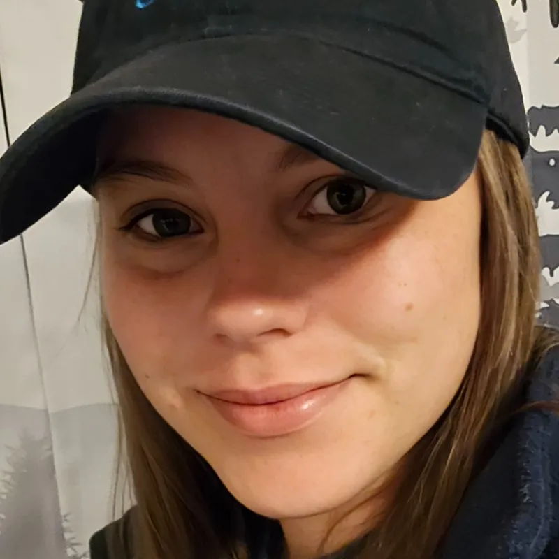 Close-up of a woman smiling wearing a black hat with a biolm logo and casual dark clothing indoors.