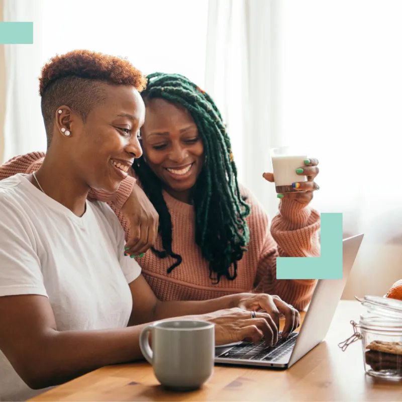 Two women smiling and working together on a laptop at a wooden table with snacks and drinks nearby