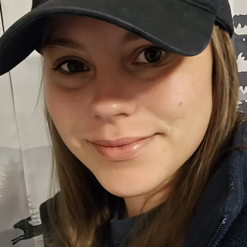 Close-up of a woman smiling wearing a black Ogiabloom logo cap and dark jacket indoors.