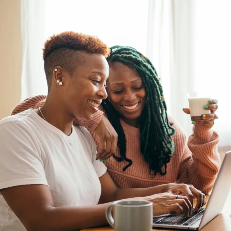 Two smiling women using a laptop together at a table with a coffee mug and a glass of milk in a bright room.