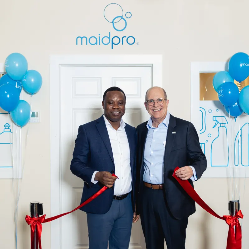 Two men in suits smiling and cutting a red ribbon at a MaidPro office event with blue balloons and cleaning graphics.