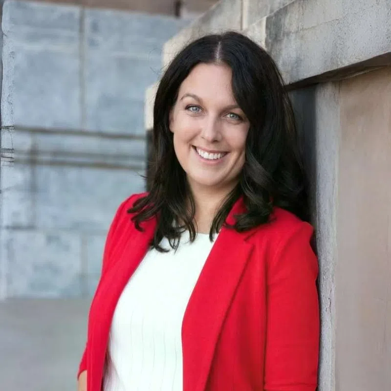 Smiling woman with dark hair wearing a red blazer and white dress leaning against a stone wall outdoors.