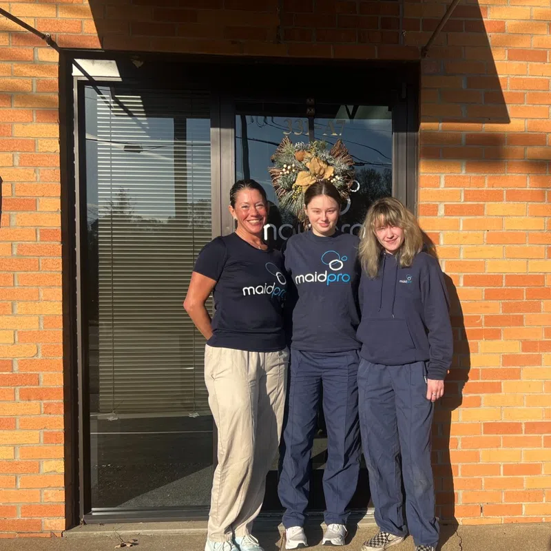 Three women in MaidPro uniforms standing outside a brick building entrance with a decorative wreath on the door.