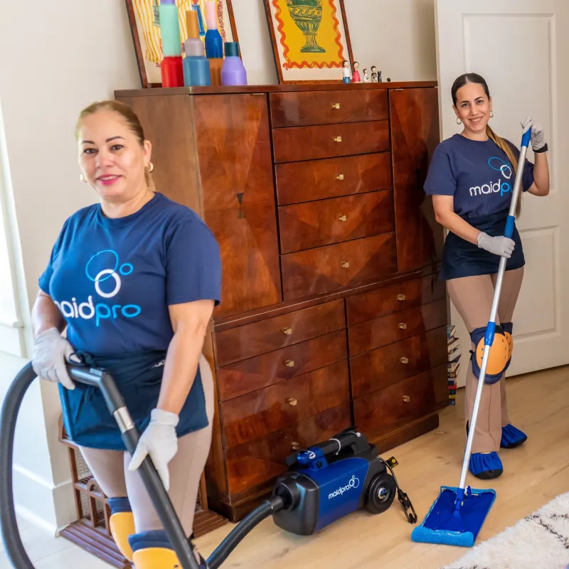Two MaidPro cleaners vacuuming and mopping a wooden floor in a well-lit room with a wooden dresser.