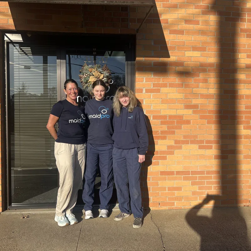 Three women in MaidPro uniforms standing outside a brick building entrance with a holiday wreath on the glass door