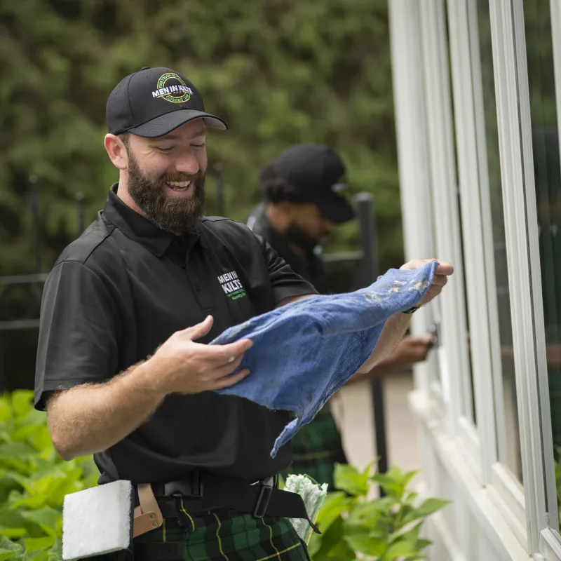 Smiling window cleaner wearing a black cap and uniform holding a blue cloth outside a house with greenery background.
